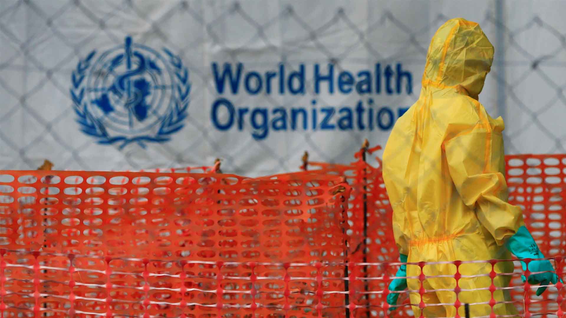 Person with hazmat suit walking in front of a WHO logo. 