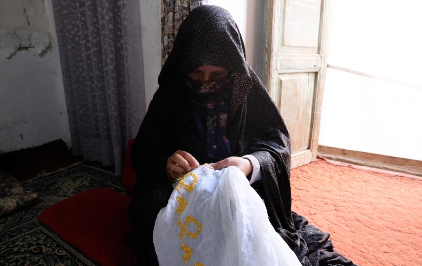 Pictured is a woman sat down and sewing a pattern with yellow thread onto a white fabric. She is wearing a polka-dot head-covering and flower-pattern clothes. 